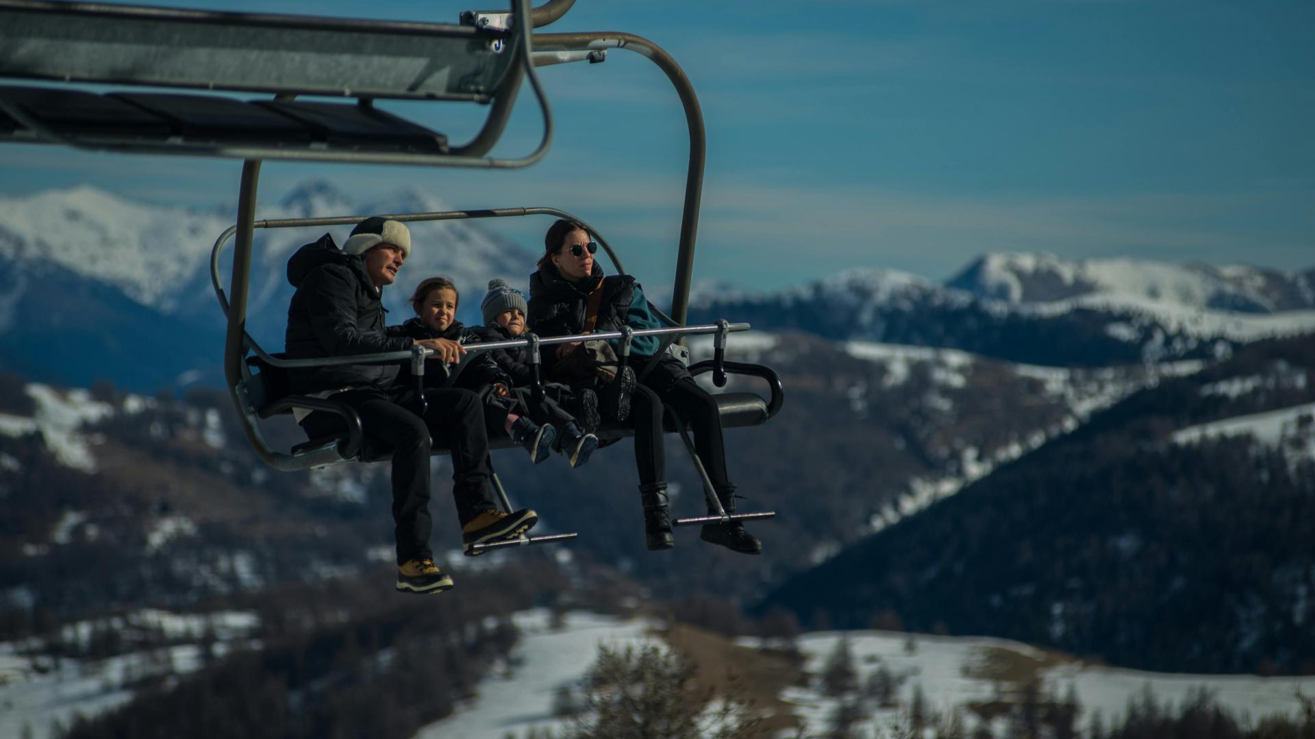 Skiers on a chairlift over snowy mountains under a blue sky.